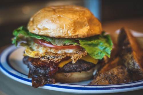A juicy burger with lettuce, tomato, cheese, and a fried egg, served on a toasted bun with crispy potato wedges.