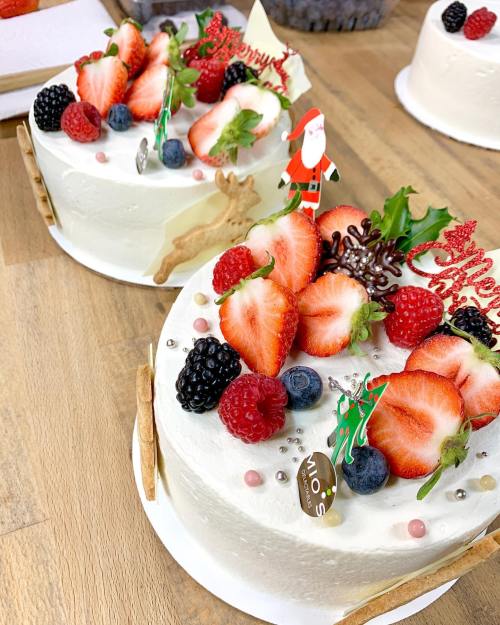 Two festive cakes topped with strawberries, raspberries, blackberries, and decorative holiday elements on a wooden table.