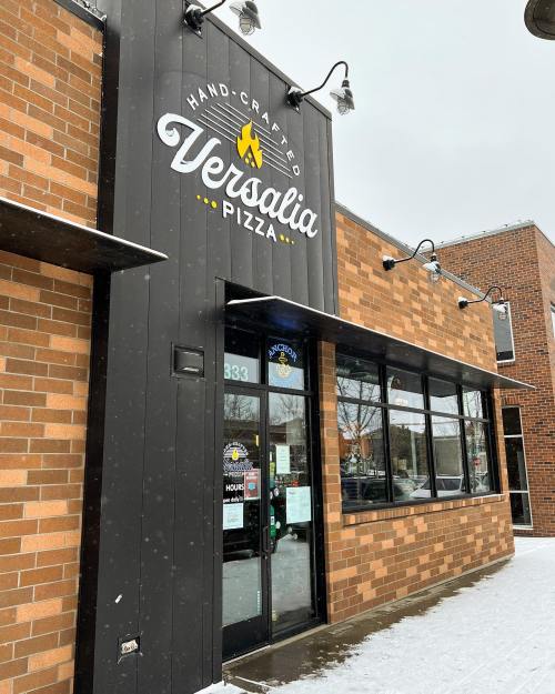 Exterior of Versalia Pizza, featuring a brick facade and large windows, with snow falling outside.