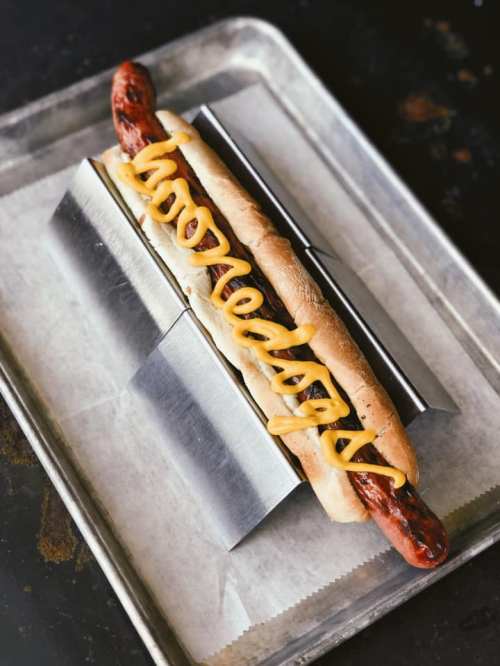 A grilled hot dog in a bun, topped with yellow mustard, served on a metal tray.