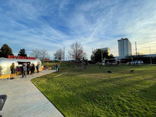 A park scene with people gathered near a food truck, dogs playing, and a skyline in the background under a blue sky.