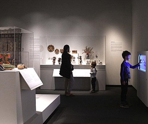 A museum exhibit featuring artifacts, with two visitors exploring and a child interacting with a touchscreen display.