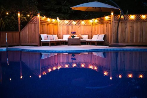 Cozy outdoor seating area by a pool, illuminated by warm lights, with a wooden fence and an umbrella.