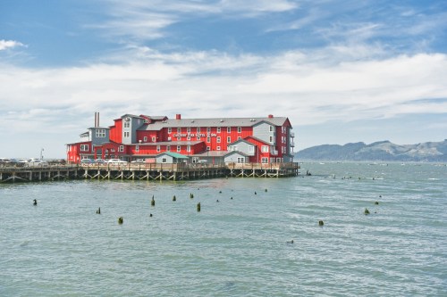A red building on stilts by the water, with mountains in the background and a cloudy sky above.