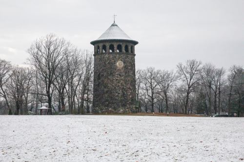 A stone tower with a conical roof stands in a snowy landscape, surrounded by bare trees under a gray sky.
