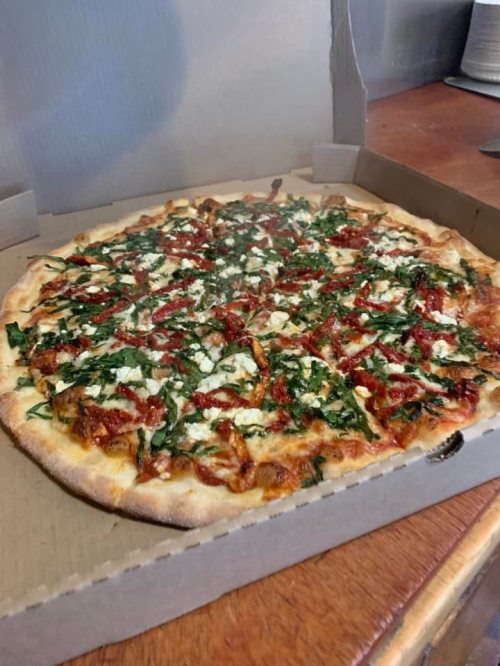 A freshly baked pizza in a cardboard box, topped with spinach, sun-dried tomatoes, and feta cheese.