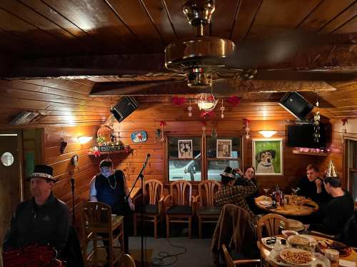 A cozy restaurant interior with wooden walls, tables, and chairs, featuring decorations and a small stage for performances.