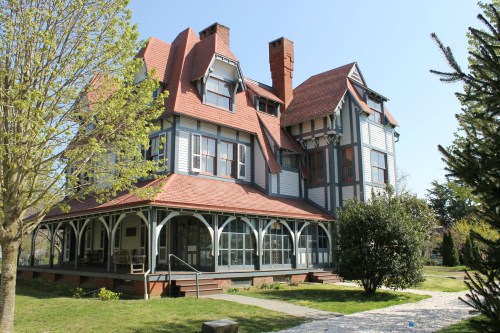 A large, historic Victorian-style house with a red roof, surrounded by greenery and a clear blue sky.