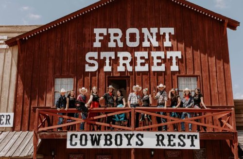 A group of people in cowboy hats poses on a balcony of a rustic building labeled "Front Street Cowboys Rest."