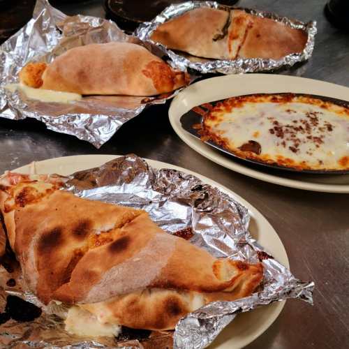 A plate with calzones wrapped in foil and a baked dish with melted cheese, all on a stainless steel surface.