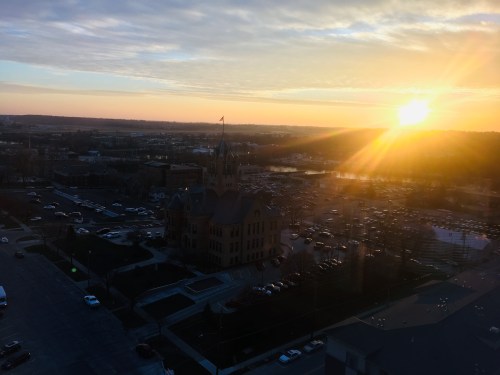Sunset over a historic building with a flag, surrounded by a parking lot and fields in the background.