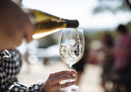A hand pours sparkling wine into a glass with a logo, set against a blurred outdoor background.