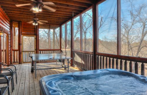 A screened porch with a hot tub, wooden flooring, and a table, surrounded by trees and natural light.