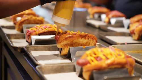 A close-up of hot dogs being prepared, topped with mustard and cheese, on metal trays in a food preparation area.