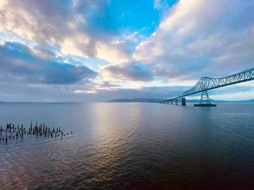 A bridge spans over calm waters, with clouds reflecting in the sky and remnants of old pilings visible in the foreground.