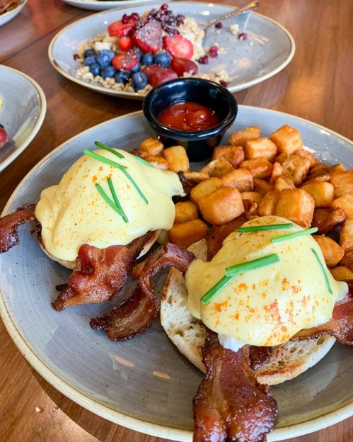 A plate of eggs Benedict with bacon on an English muffin, served with crispy potatoes and a side of ketchup.