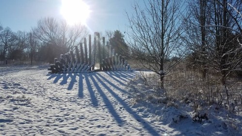 A snowy landscape features a large wooden sculpture casting long shadows under a bright sun. Trees are visible in the background.