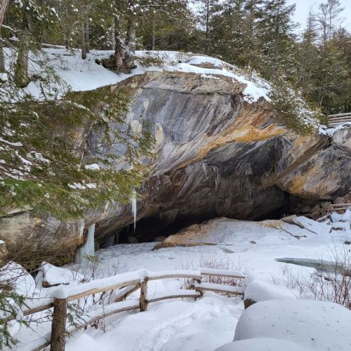 A snowy landscape featuring a rocky overhang and trees, with a wooden railing in the foreground.