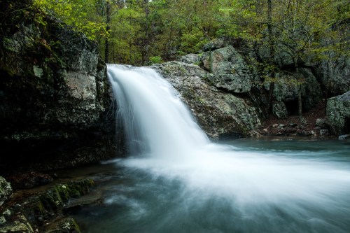 A serene waterfall cascades over rocks into a tranquil pool, surrounded by lush green trees and natural scenery.