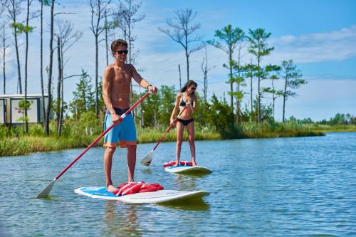 Two people paddleboarding on a calm waterway, surrounded by trees and blue skies. One is wearing a bikini, the other shorts.
