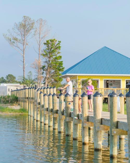 Three people stand on a wooden pier by the water, with a yellow building and trees in the background under a clear sky.
