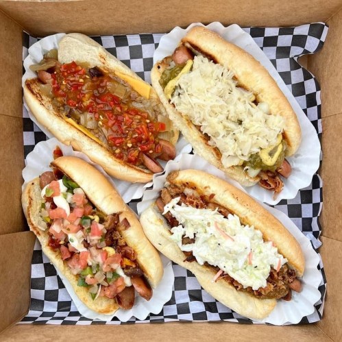 Four gourmet hot dogs in a takeout box, each topped with various ingredients like sauerkraut, bacon, and salsa.