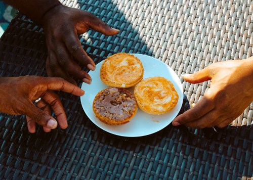 Two hands reaching for three colorful pastries on a white plate, set on a textured black surface.