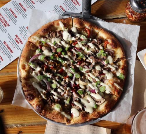 A close-up of a pizza topped with meat, vegetables, and drizzled with sauce, served on a wooden table.