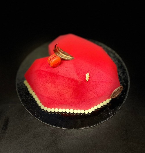 A heart-shaped red cake adorned with a macaron, a leaf, and gold accents on a black background.