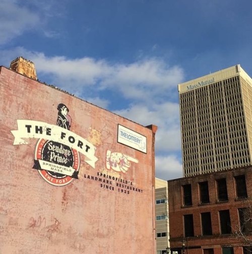 Mural of "The Fort" restaurant on a brick wall with a blue sky and Miss Mutual building in the background.