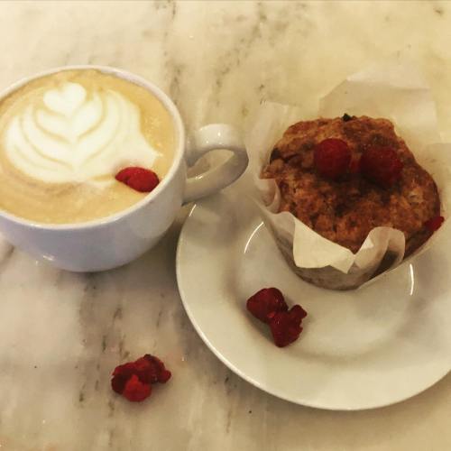 A cup of latte with latte art beside a muffin topped with raspberries, on a marble surface.