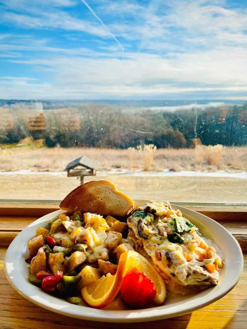 A plate of omelette, hash browns, toast, and fruit, with a scenic view of a landscape and blue sky in the background.