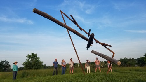 A large abstract sculpture made of logs and metal stands in a grassy field, with several people observing it nearby.