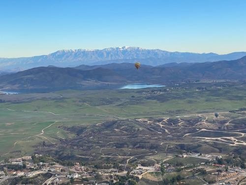 A hot air balloon floats above a scenic landscape with mountains, a lake, and green fields under a clear blue sky.