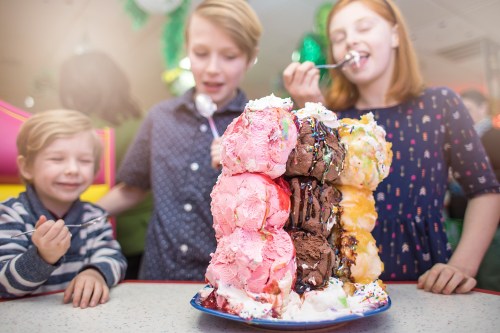Three children enjoy a towering ice cream dessert, smiling and taking bites, with colorful scoops and toppings.