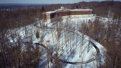 Aerial view of a snow-covered landscape featuring a large, abandoned building surrounded by bare trees.