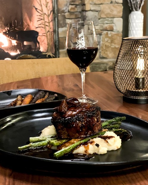 A gourmet meal featuring steak, asparagus, and mashed potatoes, with a glass of red wine and a cozy fireplace in the background.