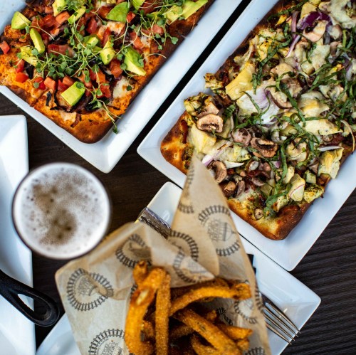 Two pizzas topped with vegetables, alongside a basket of fries and a glass of beer on a wooden table.