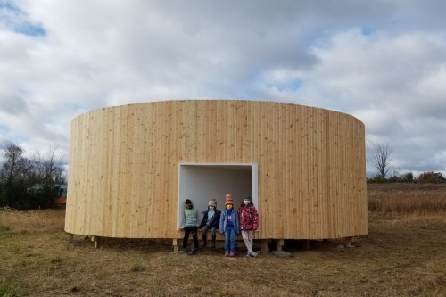 A circular wooden structure in a field, with five children standing at its entrance, under a cloudy sky.