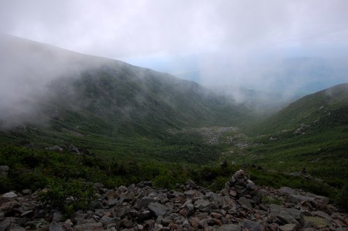 A misty mountain valley with rocky terrain and lush greenery, partially obscured by clouds.