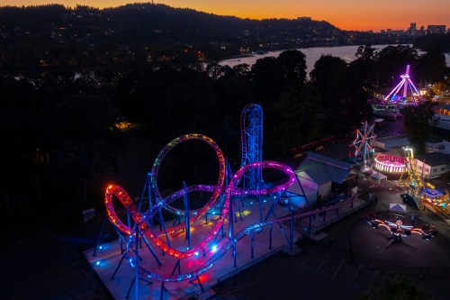 Aerial view of a colorful amusement park at dusk, featuring a lit roller coaster and carnival rides by a river.