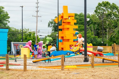 Colorful outdoor play area with oversized toys, a giant stacking game, and children playing near cars and structures.