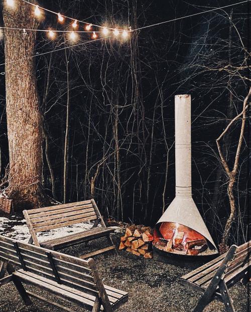 Cozy outdoor seating area with a fire pit, surrounded by trees and string lights at night.