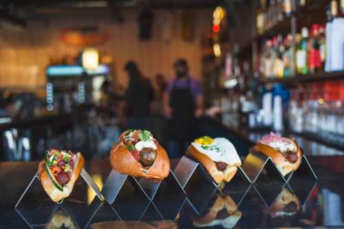 Four gourmet hot dogs on a bar counter, each topped with various fresh ingredients, with a blurred background of a bar.