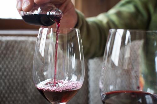 A hand pours red wine from a small bottle into a glass, with another glass partially visible in the background.
