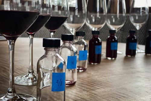 A row of wine glasses and small bottles with blue labels, filled with red liquid, arranged on a wooden table.