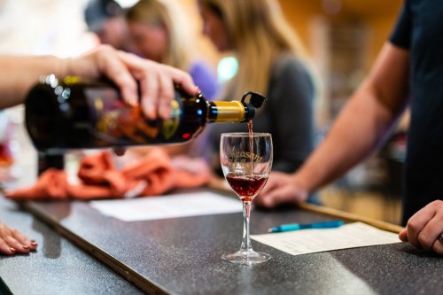 A person pours red wine into a glass at a bar, with others socializing in the background.