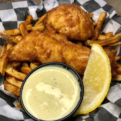 A basket of golden fried fish fillets and crispy fries, served with a lemon wedge and a small cup of dipping sauce.