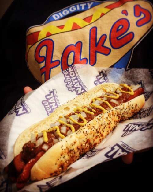 A close-up of a hot dog in a bun topped with mustard and onions, held by a person wearing a "Jake's" shirt.
