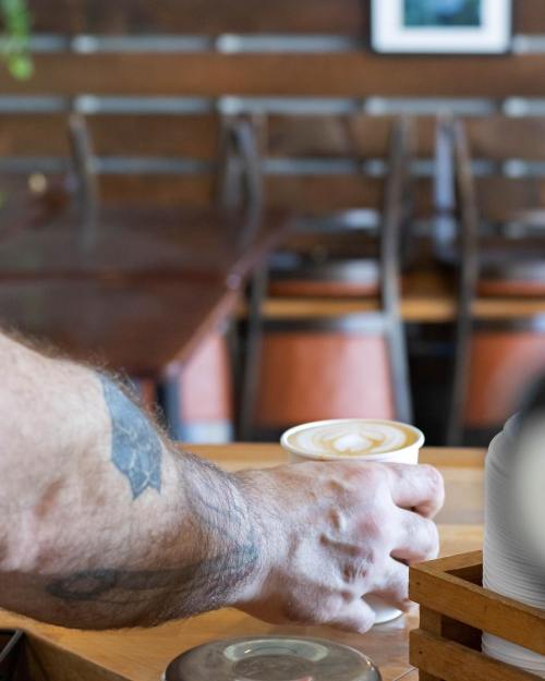 A hand with a tattoo holds a coffee cup with a heart-shaped latte art, set in a cozy café environment.